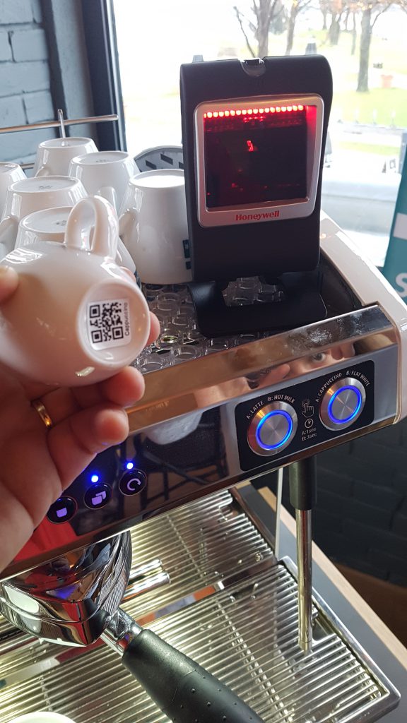 Digitale Kaffeetasse mit QR-Code unterhalb der Tasse. Scanner an Kaffeemaschine zum Erfassen der produzierten Artikel.